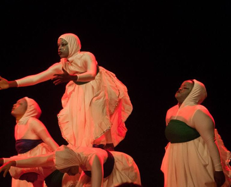 A dancer in white reaches forward over a kneeling dancer